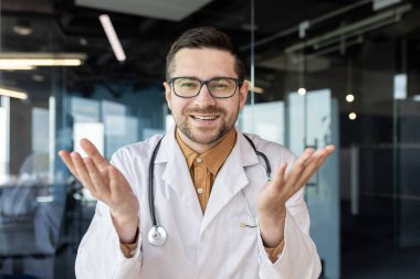 Online doktor danışmanlığı, genç başarılı doktor web kamerasına bakıyor ve müşteriyle neşeli bir şekilde gülümsüyor. Modern klinik ofisinde steteskopla uzaktan çalışan adam..