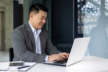 Ofisin içinde bilgisayarla çalışan başarılı olgun Asyalı, klavyede yazan ve gülümseyen adam, işten ve başarı sonuçlarından memnun iş adamı, programcı..