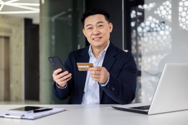 Ofisteki başarılı Asyalı adamın portresi, iş adamı kameraya bakıyor ve gülümsüyor, online alışveriş için banka kartı ve cep telefonu tutuyor..