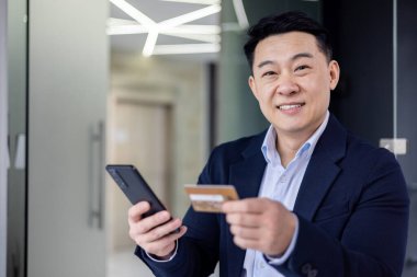 Closeup portrait of successful and satisfied asian man inside office, businessman smiling and looking at camera, mature man using money transfer app on phone, holding bank credit card.