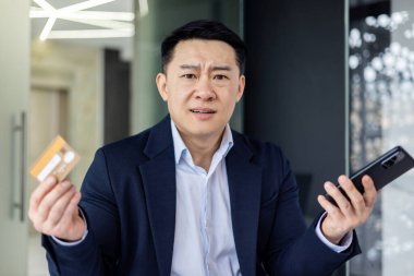 Portrait of disappointed and angry businessman inside office, cheated asian man looking at camera upset holding bank credit card and phone, man got rejection and error in online money transfer.