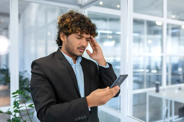 Upset hispanic man in office at work reading bad news from phone online, businessman in shirt near window working inside office, using smartphone.