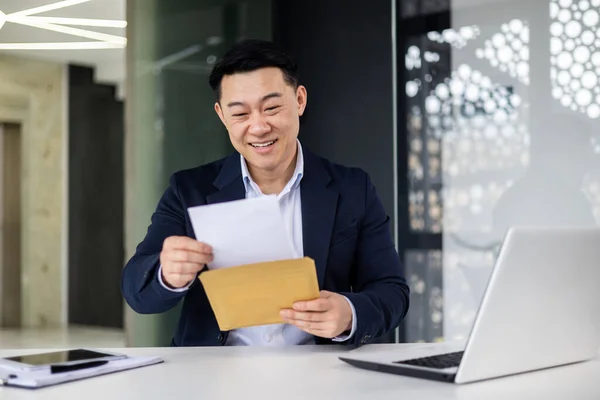 stock image Happy asian man in glasses received letter opens and reads message document, businessman works inside modern office building on laptop, good news in notification