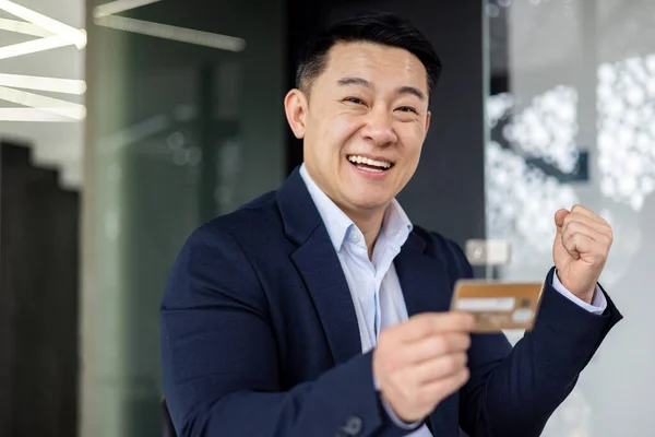 Closeup portrait of happy businessman banker inside office at workplace, man holding bank credit card in hands smiling and looking at camera, hand up gesture of victory and triumph celebrating.