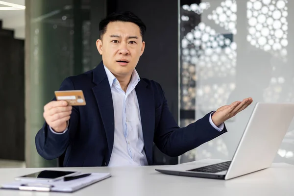 Frustrated angry asian man inside office, mature boss looking at camera and holding bank credit card, man rejected online shopping payment, businessman cheated by scammers online