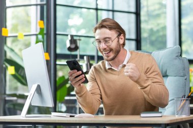 A young man works in the office at a table using a laptop. He holds the phone in his hands, looks at the screen happy and shocked, raised his glasses on his face in surprise. Got good news.