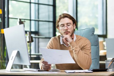 Ofis içi evrak işlerinde ciddi ve odaklanmış finansör, hesap makinesi ve dizüstü bilgisayar kullanan olgun bir adam raporları özetliyor ve günlük giysiler içindeki iş adamı