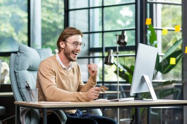 Successful online shopping. A young man in the office uses a credit card, makes orders and purchases through a laptop on the Internet. The happy man looks at the monitor, shows a yes gesture.