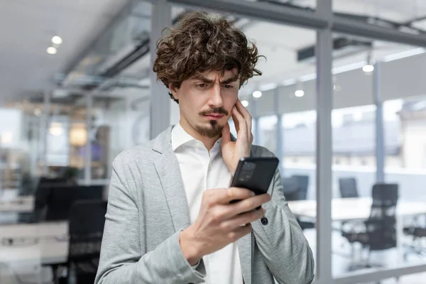 Businessman in office near window upset reading message online notification from phone, male boss working inside office in business suit with beard, upset investor.