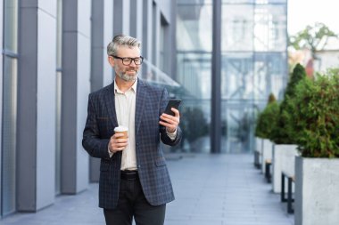 Yakışıklı bir kıdemli işadamı elinde bir fincan kahveyle bir ofis merkezinin önünde duruyor ve telefonu kullanıyor..