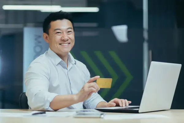 Financial transactions with a credit card. Young Asian man working in office at laptop and using credit card, posing and smiling at camera.