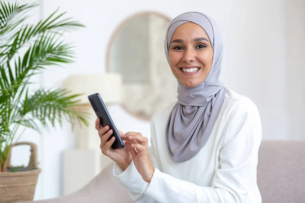 Young Muslim woman in hijab sitting on sofa at home and using phone. Smiling, she looks at the camera.