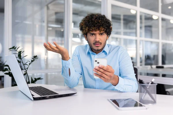 stock image Worried young hispanic man sits in the office at the desk and looks disappointedly at the phone, spreads his hands, looks upset at the camera.