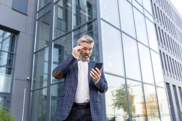 A worried senior man in a suit stands outside an office center and looks disappointedly at the phone screen, putting his glasses down in surprise with his hand on his face.