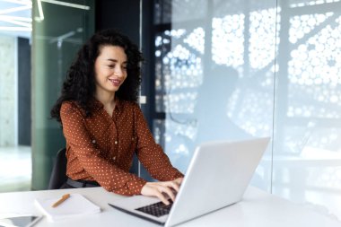 Mutlu ve gülümseyen İspanyol iş kadını dizüstü bilgisayarda yazıyor. Kıvırcık saçlı ofis çalışanı, ofis içi iş yerindeki başarılarından memnun.