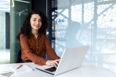 Modern ofiste çalışan genç ve güzel bir İspanyol kadın, iş kadını gülümseyen ve bilgisayarını kullanarak iş yerinde kameraya bakan.