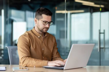 Genç bir erkek programcının portresi, kahverengi gömlekli ve gözlüklü, ofiste bir masada otururken bir dizüstü bilgisayar üzerinde çalışmaya odaklanmış..