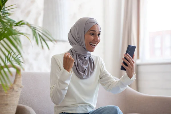 stock image Happy young Arab woman in hijab sitting on sofa at home and using mobile phone. looks at the screen and makes a victory gesture with his hand.