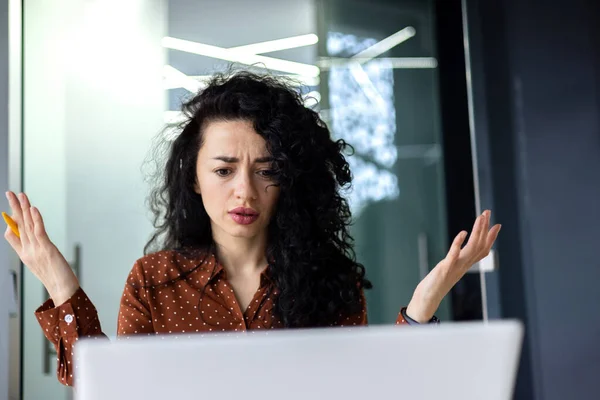 Upset and nervous business woman looking at laptop screen, female worker working inside office building, frustrated and dissatisfied with result, spreading hands.