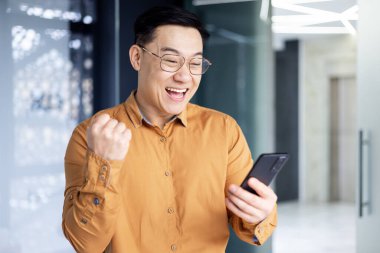 Successful asian boss near window received good news online, businessman use phone and celebrate victory and good achievement results inside office, man in shirt and glasses excitedly rejoice