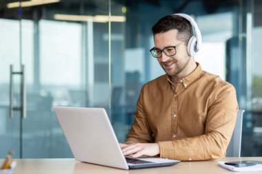 Ofiste oturan, kulaklık takan ve internette dizüstü bilgisayar kullanan gülümseyen bir erkek öğrencinin portresi..