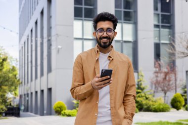 Hintli işadamının portresi, telefonu tutan, gülümseyen ve kameraya bakan bir adam, modern ofis binasının dışında gömlekli bir ofis çalışanı, öğle yemeği molasında..