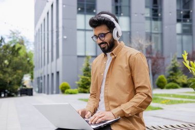 Genç başarılı Hindu öğrenci dizüstü bilgisayar kullanıyor, kulaklık takıyor müzik dinliyor, internet kitapları dinliyor, ses yayını yapıyor, klavyede daktilo yazıyor, üniversite kampüsünün yanındaki bankta oturuyor..