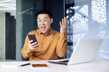 Successful and happy asian businessman man celebrating victory sitting and working in modern office at desk, celebrating victory looking at camera and joyfully shouting holding mobile phone.