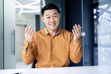 Çevrimiçi webinar. Aktif bir Asyalı genç iş adamının portresi. Kamerada internetten bir web sitesi yönetiyor, modern bir ofiste bir masaya oturuyor, konuşuyor, anlatıyor, elleriyle işaret ediyor, gülümsüyor.