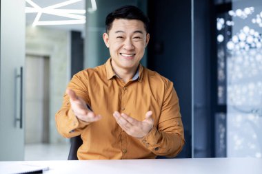 Successful and happy Asian businessman looking at camera, talking on video call, cheerfully gesticulating to interlocutor, online meeting, man working in office at work.
