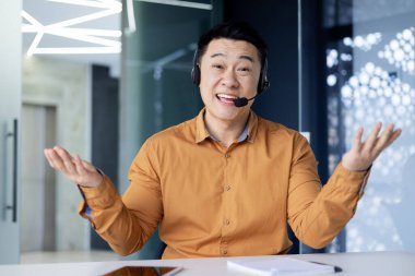 Cheerful and smiling online customer support tech worker, Asian man with call headset smiling and looking at webcam, working on helpline and consulting customers, using headset