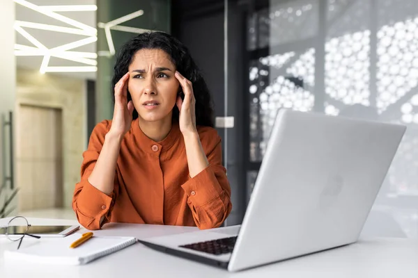 stock image Beautiful woman at workplace overworked, tired hispanic woman at workplace with severe headache, businesswoman working inside office sitting with laptop, stressed and sad.