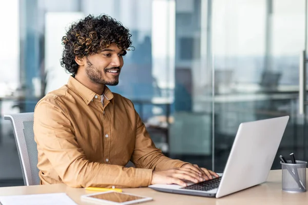 Genç, çağdaş iş adamının portresi ofis içi, işyerinde gülümseyen bir adam, genç İspanyol iş adamı dizüstü bilgisayar kullanarak masada oturuyor.