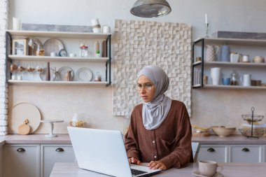 Uzaktan kumanda. Tesettürlü genç Müslüman bir kadın dizüstü bilgisayarla evde çalışıyor. Mutfakta, masada oturmuş, endişeli ve şaşkın bir şekilde monitöre bakıyorsun..