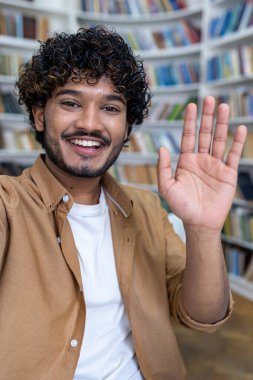 Genç, mutlu bir İspanyol erkek öğrenci kütüphanedeki bir telefondan video görüşmesi yapıyor. Kameraya selamlar ve dalgalar, uluslararası öğrenci değişim programına katılıyor. Dikey fotoğraf.