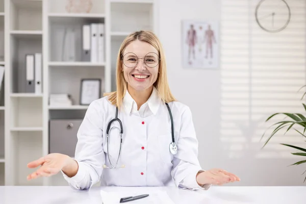一名女医生身穿白衣 带着听诊器坐在桌旁办公室里 在网上用摄像机交谈和咨询的画像 他笑着 主持医学网络研讨会 — 图库照片