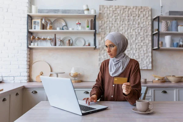 A Muslim young woman wearing a hijab is sitting in the kitchen using a laptop and holding a credit card. Makes online purchases, pays bills, transfers funds, checks the budget.