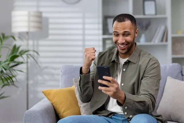 stock image Young smiling Muslim man using phone while sitting at home on sofa, playing online games, placing bets, enjoying money win, celebrating, showing victory gesture with hand.