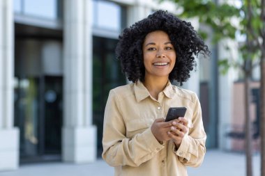 Kampüsün dışında elinde telefonla yürüyen, iş kadını gömleğiyle gülümseyen ve mesaj yazarken kameraya bakan güzel Afro-Amerikan kız öğrencinin portresi..