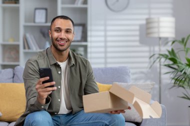 Evinde, elinde telefon, elinde bir paketle kanepede oturan mutlu bir Afro-Amerikalı 'nın portresi..