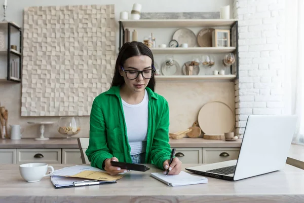 stock image A young woman works at home in the kitchen with a laptop and documents. A housewife deals with financial family issues and the budget, uses a calculator.