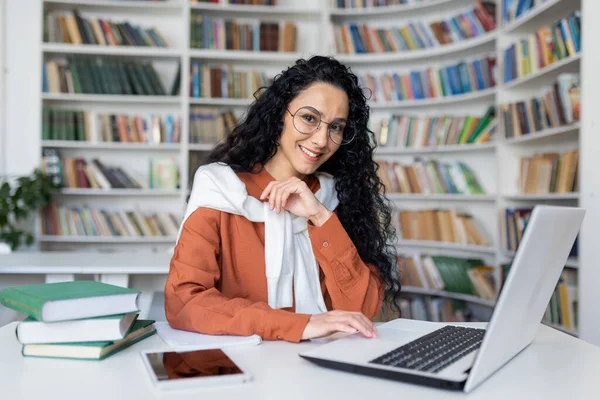 Young hispanic woman studying in academic university library, female student smiling and looking at camera while sitting at laptop, woman with curly hair.