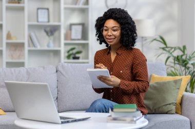 Mesafe öğrenme. Genç bir Afrikalı Amerikalı kız öğrenci evdeki kanepede oturuyor ve bir dizüstü bilgisayar kullanarak internetten ders çalışıyor. Not alır ve konferansı bir defterde dinler..