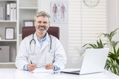 Beyaz önlüklü, steteskoplu, gülümseyen ve kameraya bakan yaşlı, gri saçlı bir erkek doktorun portresi..