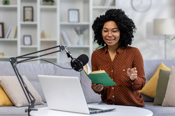 A young African-American woman journalist, blogger conducts a podcast, broadcast remotely from home. Sitting on a sofa in front of a microphone and a laptop and reading a book.