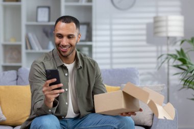 Tatmin olmuş Latin Amerikalı adam evinde kanepede oturuyor ve elinde tutuyor. Eve kuryeyle getirdiği paketi açıyor. Telefonu kullanıyor, uygulamayla online sipariş veriyor..
