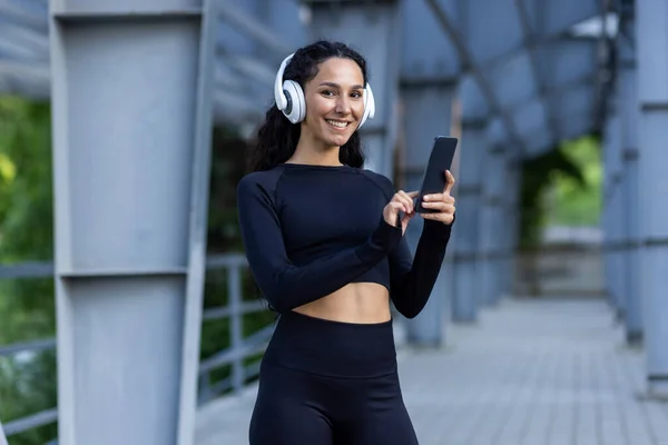 stock image Portrait of happy and successful hispanic woman, sportswoman in headphones and tracksuit looking at camera, woman using online app for listening to music, and exercising outdoors.