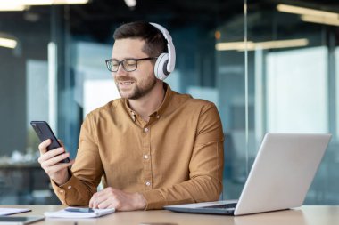 Kulaklıklı, elinde telefon olan gülümseyen genç bir adam ofisteki bir masada oturuyordu. Çalışıyor, dinleniyor, video görüşmesinde konuşuyor, müzik dinliyor..