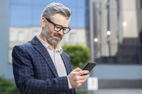 A successful gray-haired businessman uses phone outside an office building, a senior boss in a business suit walks down the street, a man reads messages and browses online pages, uses an application.