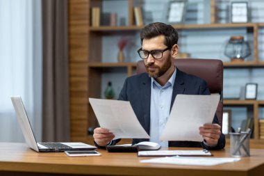 Ofis içi evrak işlerinde ciddi ve odaklanmış finans danışmanı, hesap makinesi ve dizüstü bilgisayar kullanan olgun bir adam raporları ve hesapları özetliyor, iş adamı.